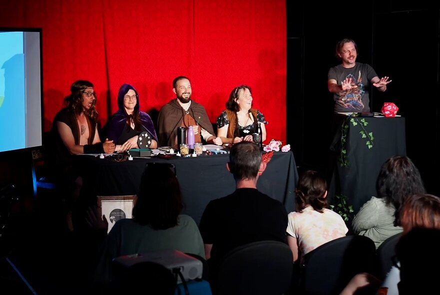 Four performers seated at a table on a stage behind a table, with a person guiding them through a Dungeons and Dragons style improvised adventure. 