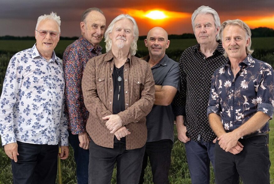 Country rock band Stars six-piece line-up of older musicians wearing country shirts standing in front of a sunset sky.