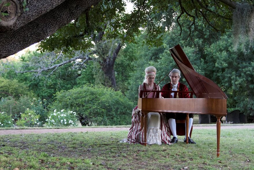 Two people wearing 18th century clothing sitting in front of a grand piano and playing, outdoors in a large garden.