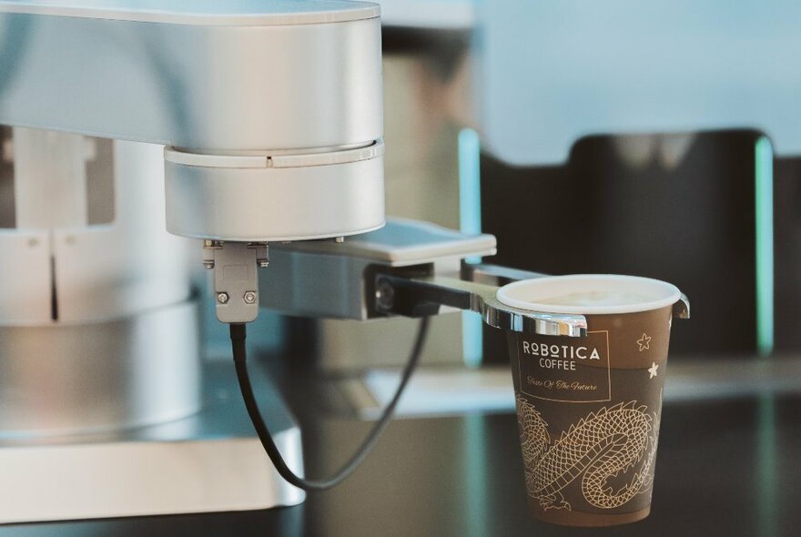 A robot barista holding a brown takeaway cup of coffee in metal tongs.