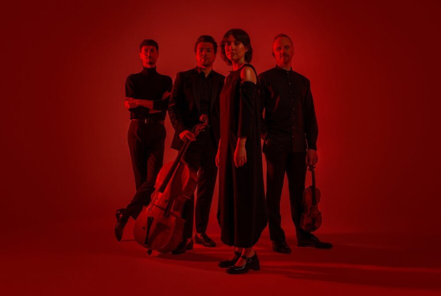 The four musicians of the Australian String Quartet, posed standing together in a group, some holding their instruments, with a dark red filter over the image.