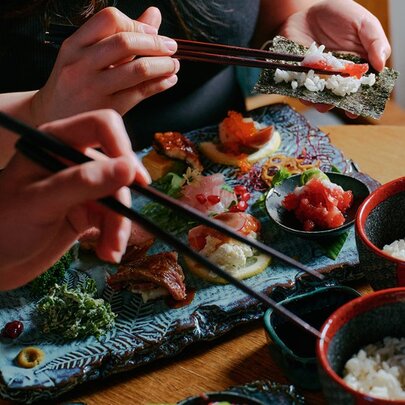 Hands holding chopsticks taking rice and items from bowls and large sushi platter.