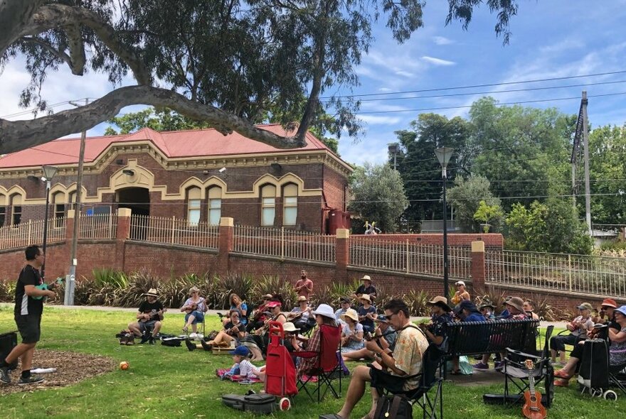 Group of people seated outdoors on a grassy area with many of them playing ukuleles, a building visible behind them.