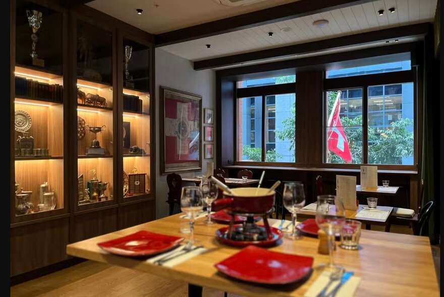 A table in small restaurant set with red plates and a fondue pot,  the Swiss flag visible through a window.