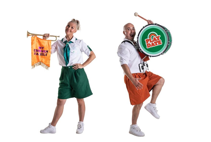 Two people wearing baggy shorts and white shirts, one banging a drum and the other holding a trumpet.