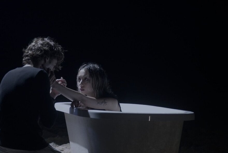 A person in a white bathtub with this arm reaching out to touch the face of a person crouched next to the bathtub next to them.
