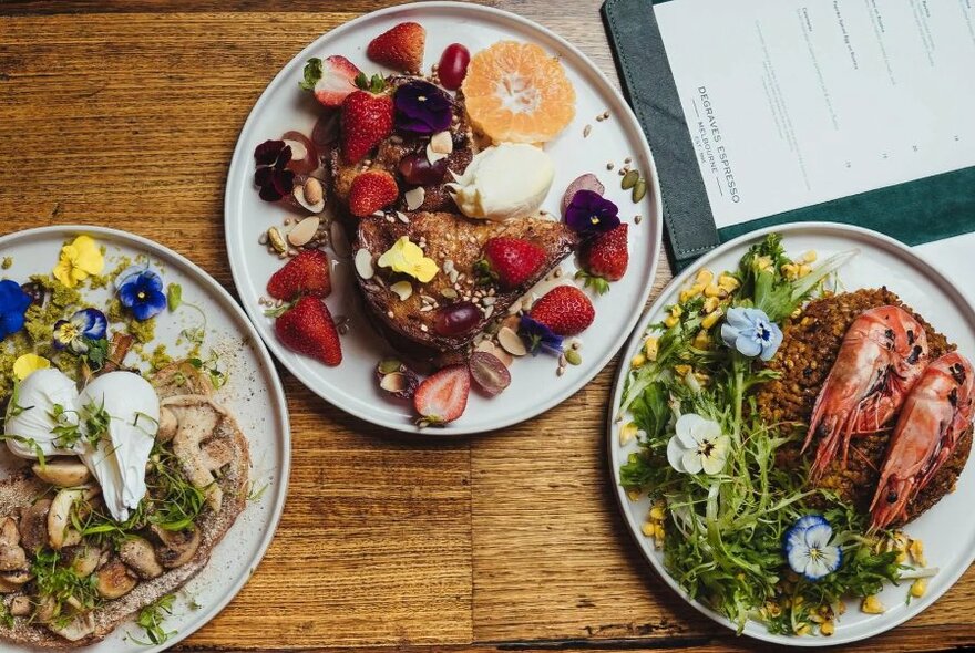 Overhead view of three plates of delicately arranged food on white plates, with a menu partially visible in the top right corner.