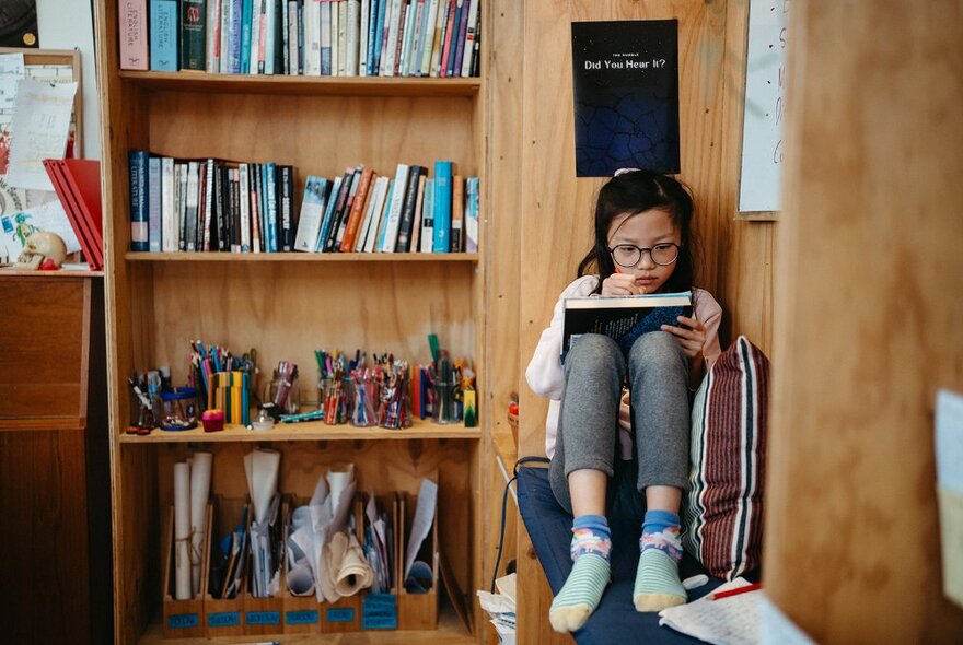 A child seated on a cushion in a study nook and writing in a workbook, next to a bookshelf, inside a writing studio space.