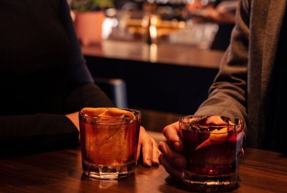 Two cocktails in glasses on a table, with two arms gently resting on the table near the drinks and close together, with a blurred bar in the background.