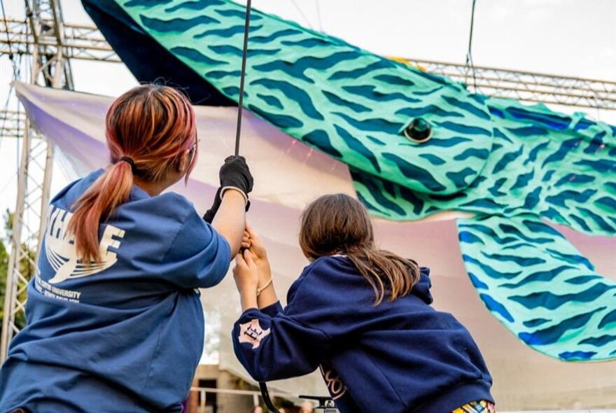 A professional puppeteer and a young person pull on a rope together, to animate a 10-metre long blue patterned whale puppet.