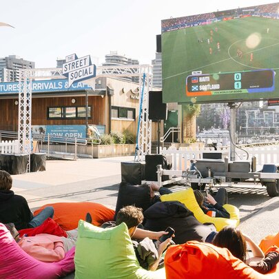 An outdoor event space at Harbour Esplanade Docklands, featuring a large screen broadcasting a sports match to an audience seated in colorful bean bags.