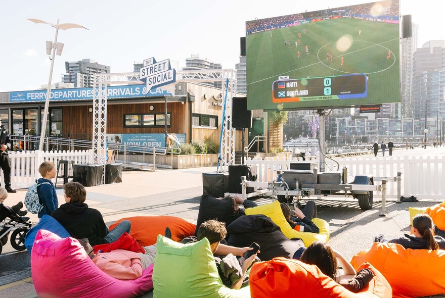 An outdoor event space at Harbour Esplanade Docklands, featuring a large screen broadcasting a sports match to an audience seated in colorful bean bags.