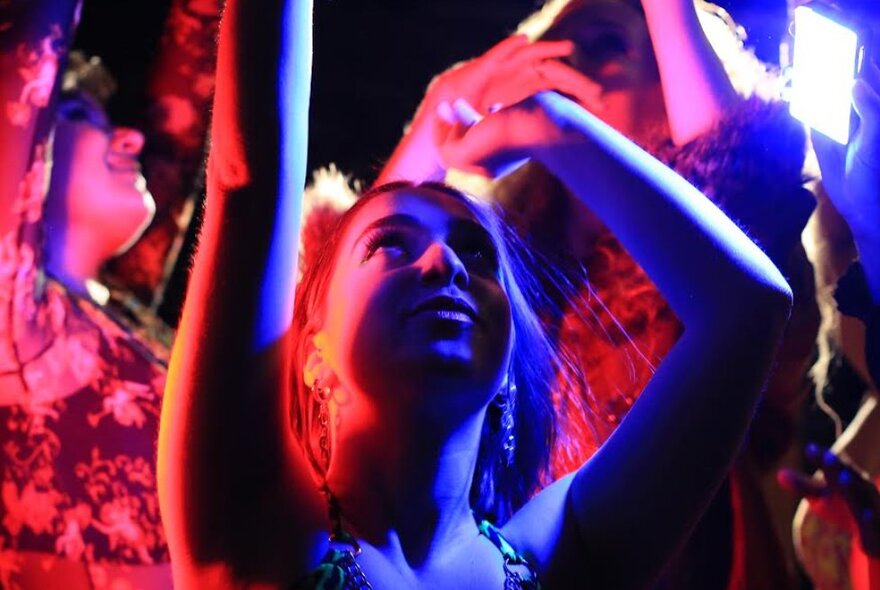 A woman at an event with her hands in the air and coloured lights cast upon her. 