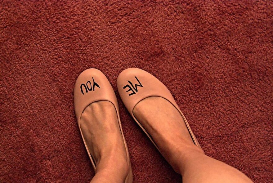 Looking down at feet wearing a pair of pale pink flat shoes and standing  on brown carpet, with the words YOU written on the top of one shoe and ME written on the other shoe.