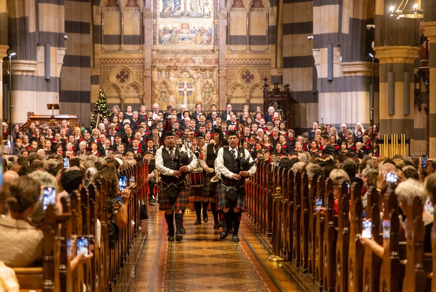Men playing bagpipes walking down the central aisle of a cathedral, with a standing choir in the background, and people seated in pews on either side of the aisle.