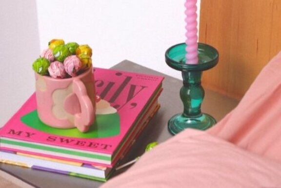 A stack of pink books on a bedside table, with a pink mug on top filled with lollipops, alongside a green candelstick and part of the bed with pink linen.
