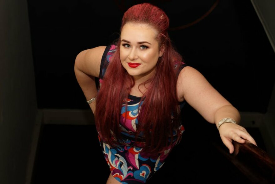 A woman with long bright red hair standing and looking up, from the bottom step of a staircase.