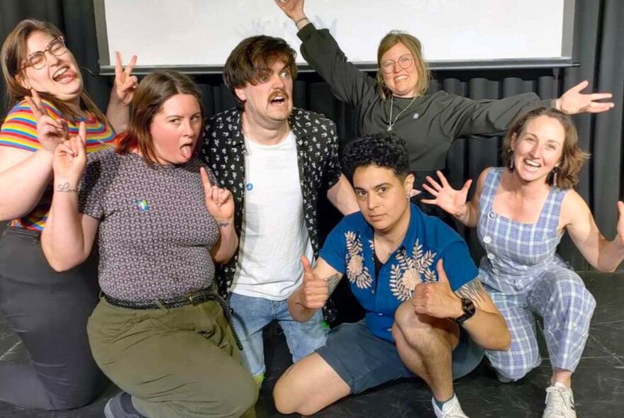 Group of people making faces and doing funny poses against a white projector screen and black wall.