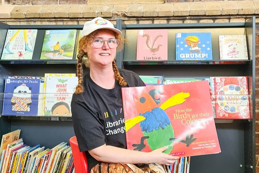 A woman with two red plaits and a white cap holds up a picture book in a kids library area. 