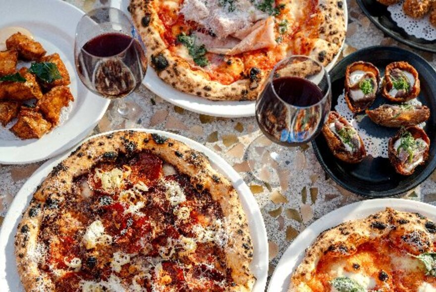 Pizzas and glasses of red wine on a restaurant table.
