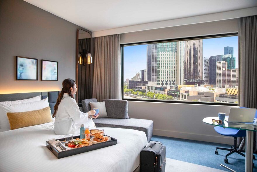 A woman in a white bathrobe sitting on a hotel bed, with a room service tray, looking at the city of Melbourne's skyline through the window.