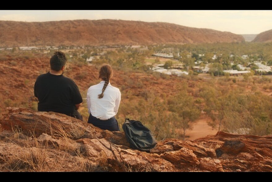 Still from the film Under Streetlights of two actors sitting on a rocky low rise hill looking at a town in the distance.