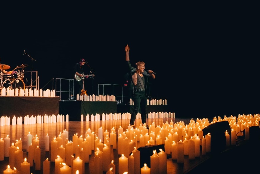A man on a candlelit stage, singing into a microphone with arm outstretched, musicians playing behind him.