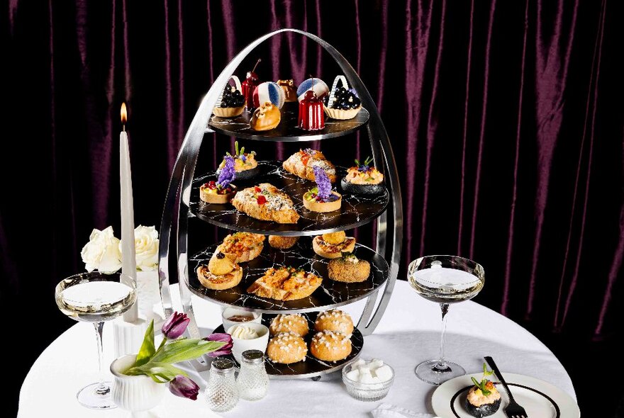 A selection of small cakes and desserts on a tiered display stand, with a glass of champagne and a lit candle on the table next to it.