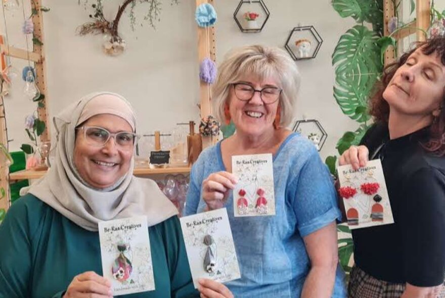Three smiling workshop participants in a studio space, holding up jewellery they have made.