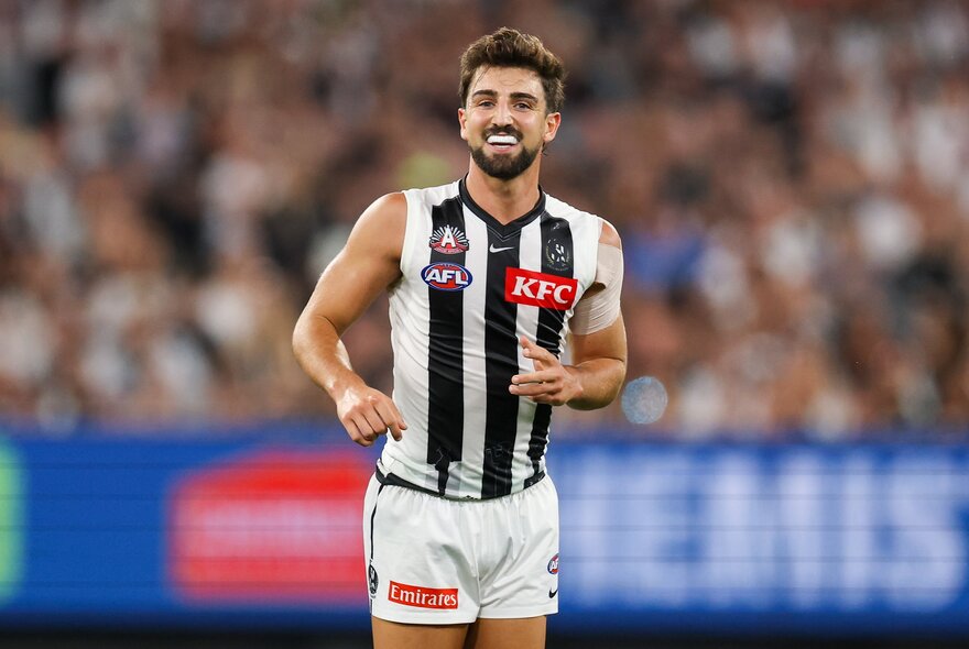 A smiling Collingwood AFL football player running on the field during a match.