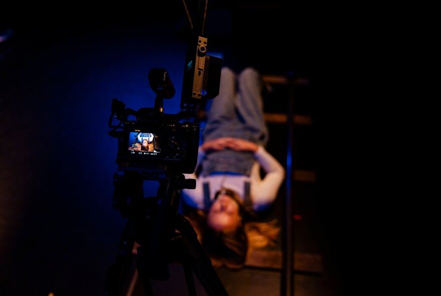 A camera seen from behind, with its digital screen in sharp focus; the subject of the photography, an 11-year old girl in overalls, lays upside down behind it, blurred.