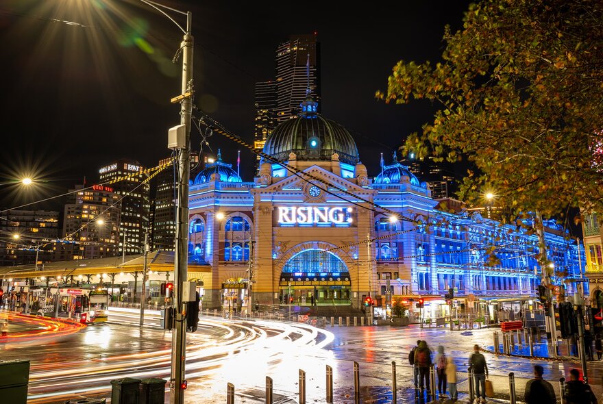 Flinders station is lit up in blue lights at night there is a big glowing sign saying rising on the front