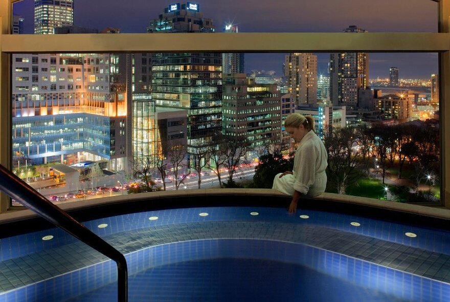 A women sitting on the edge of a spa overlooking a city skyline at night.