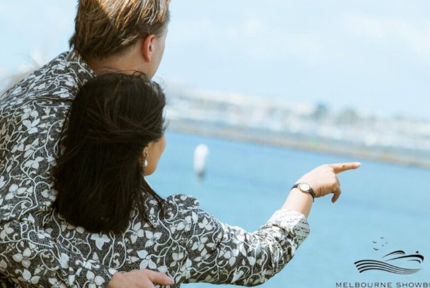 Rear view of a couple arm-in-arm standing on the deck of a boat, one of them pointing, water in the background.