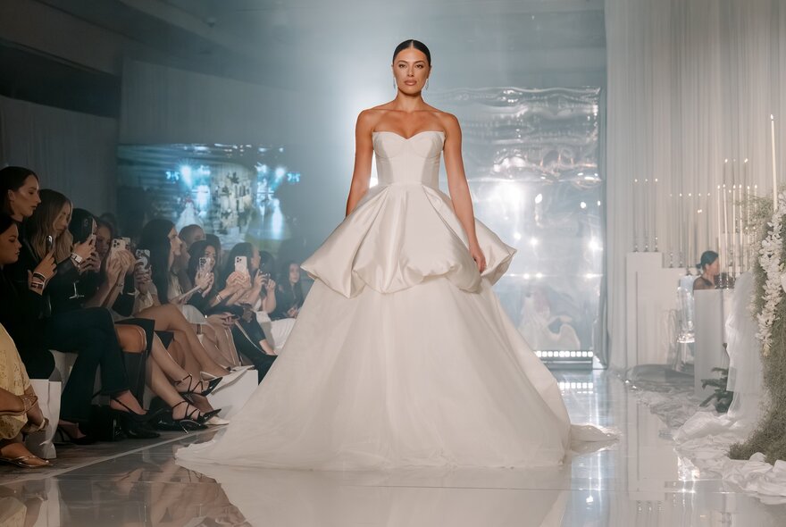 A fashion model wearing a wedding gown on a runway with a seated audience watching.
