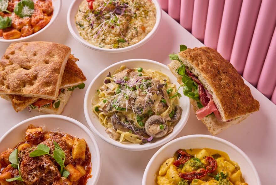 Several bowls of pasta and filled foccacia sandwiches on a table next to a pleated pink wall.