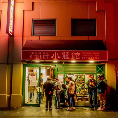 A queue of people are lining up outside a Chinese restaurant at night there is a sign saying shanghai street on the exterior of the restaurant
