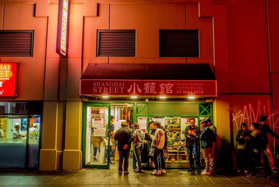A queue of people are lining up outside a Chinese restaurant at night there is a sign saying shanghai street on the exterior of the restaurant