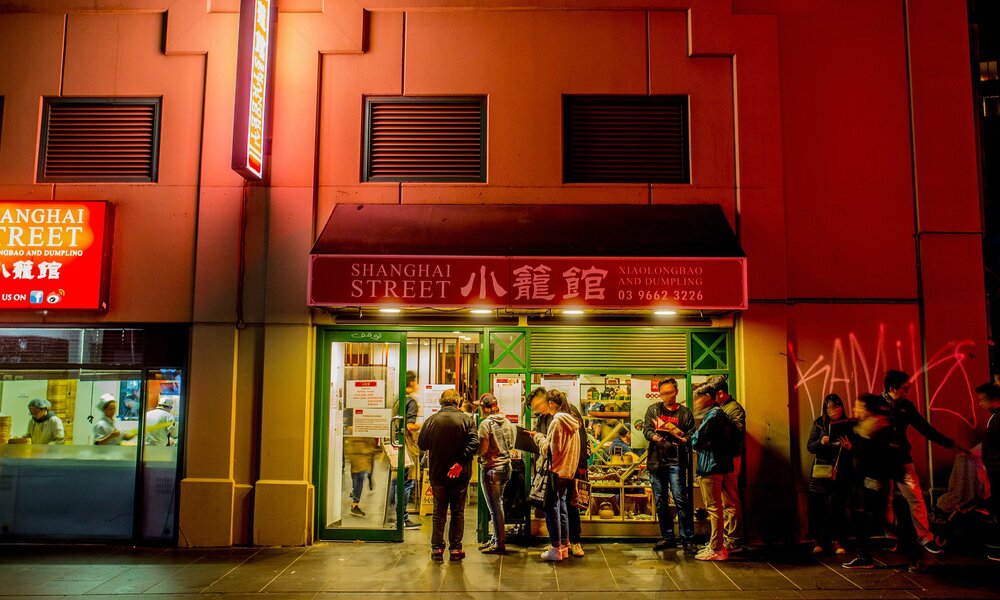 A queue of people are lining up outside a Chinese restaurant at night there is a sign saying shanghai street on the exterior of the restaurant