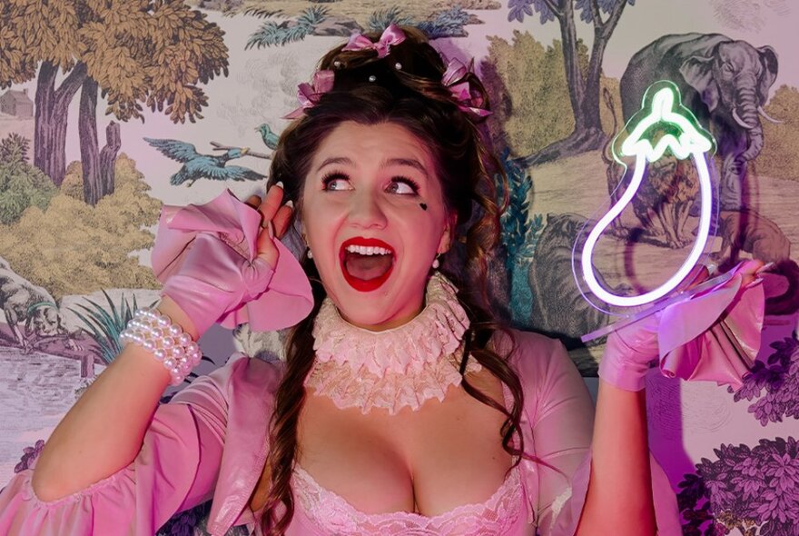 Esmé Louise James, wearing a vibrant pink outfit with ruffled sleeves, gloves, lace bodice and ruff, holding up a neon eggplant in front of pink and green patterned wallpaper.