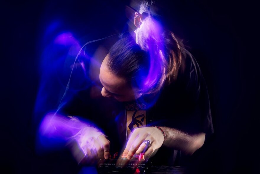 A DJ bent low over a mixing desk and twirling the controls, the image slightly out of focus due to the movement of the DJ which creates a blurred effect.