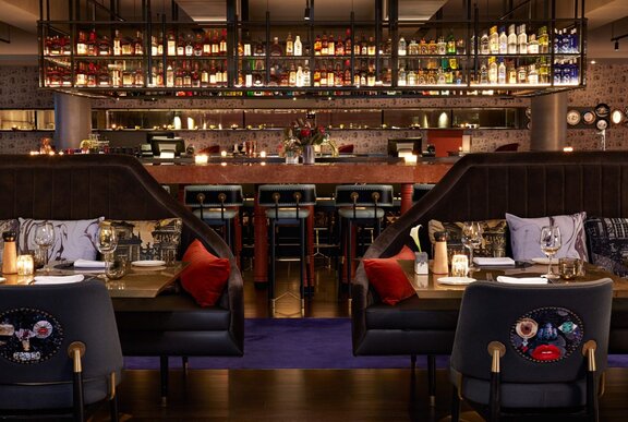 The glowing interior of a hotel bar, with bar seating and tables with embroidered velvet chairs.