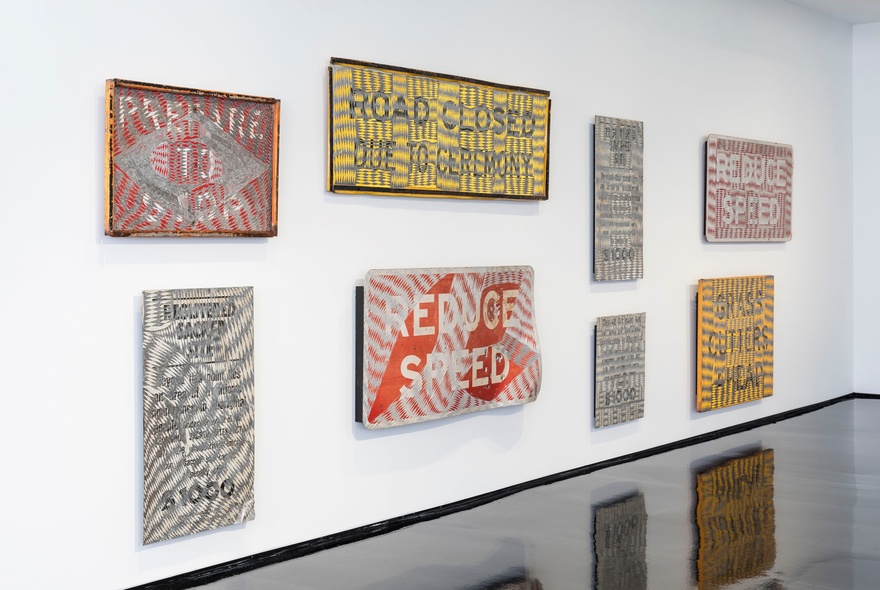 A gallery space with a white wall and shiny black floor, displaying eight pieces of artwork that look like colourful road signs.