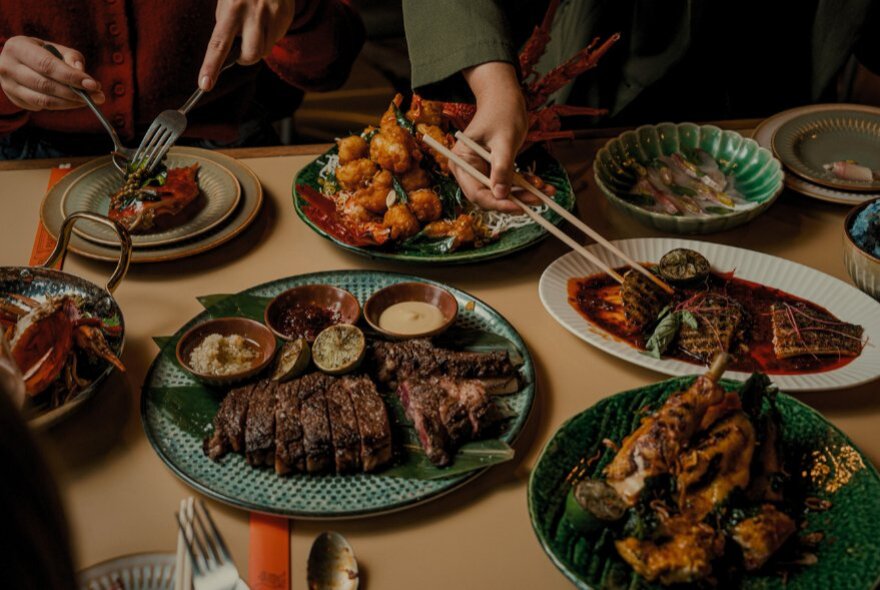 Shared platters of food on a table in a restaurant with people using forks and chopsticks to pick up food to transfer to individual plates.