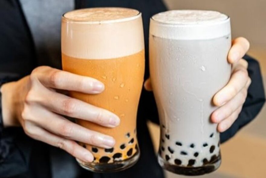 Two hands carrying two full pint glasses of bubble tea.