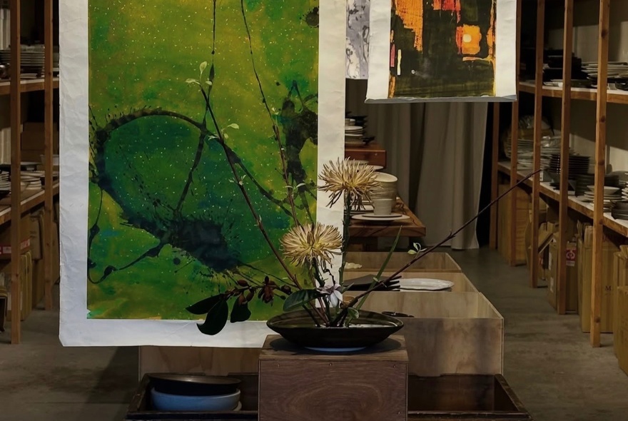 A traditional Japanese ikebana floral arrangement displayed on a wooden block in front of a green abstract artwork, in a gallery space.