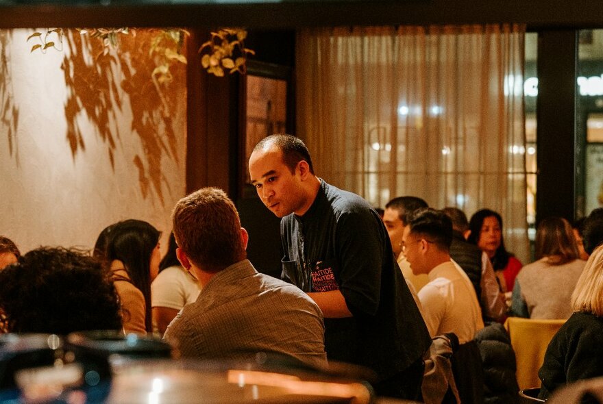 A view across a restaurant room with a waiter conferring with a diner and other diners at tables in the background