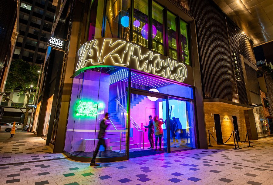 People entering a restaurant with neon lights and a sign reading 'Yakimono'.