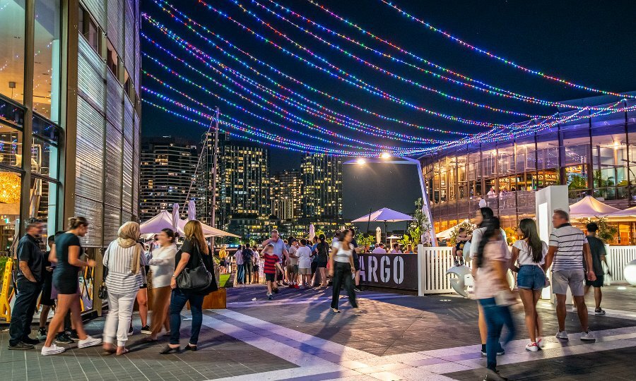 Crowds outside restaurants at night with colourful lights above them.