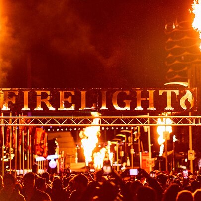 Large lettering saying FIRELIGHT above a stage at the Firelight Festival with two large flames shooting into the air at either end. 